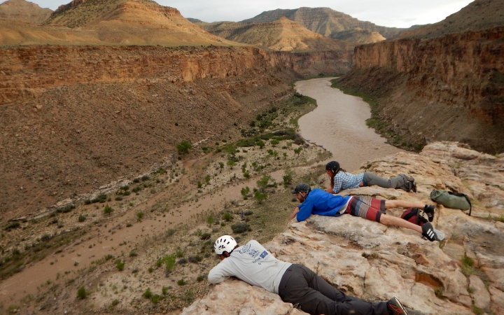Three people lay on a cliff above a river, looking down. 